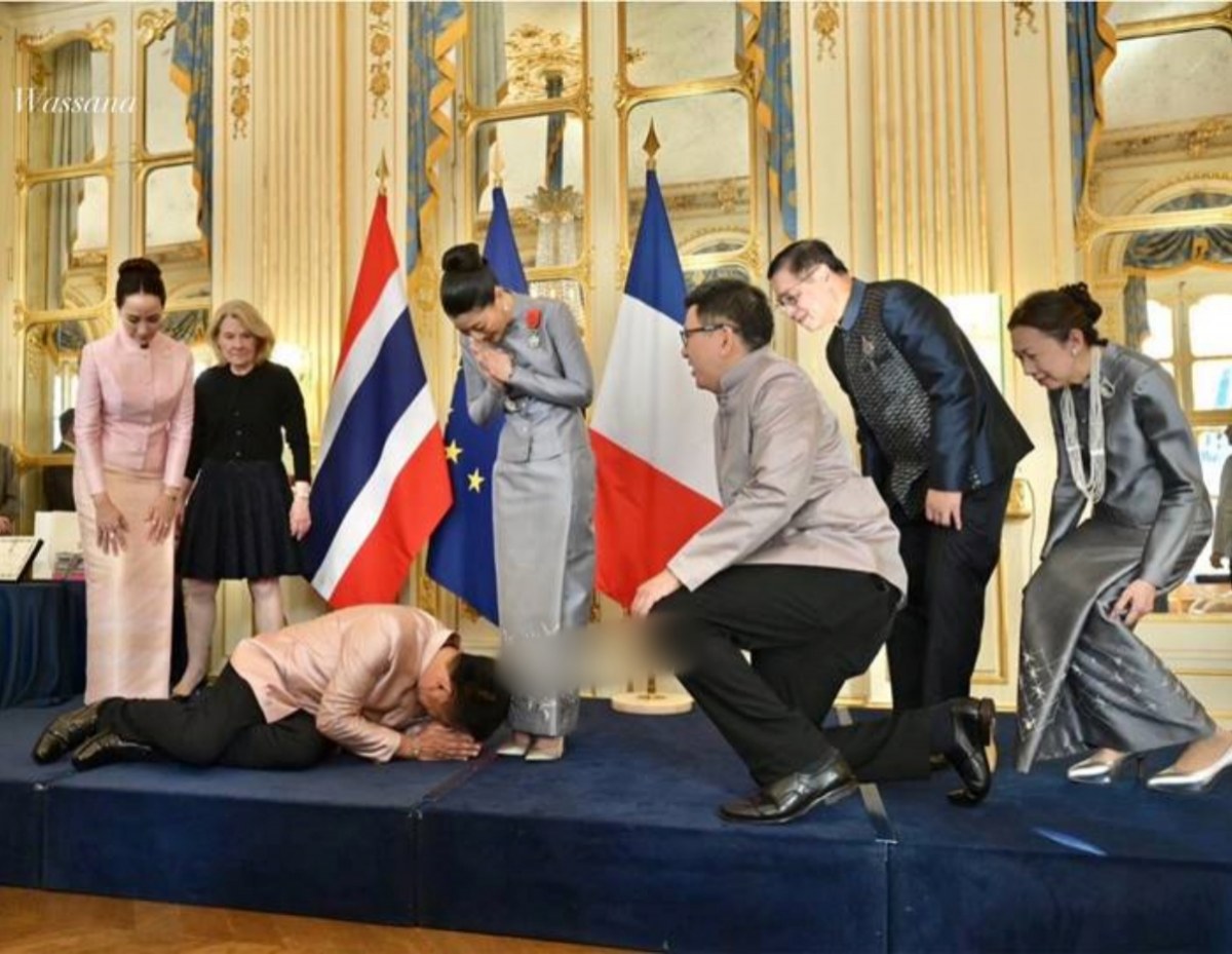 Dlock of dignitaries bowing on a blue-carpeted stage with gold ornate walls and flags (UK, EU, France) in the background.