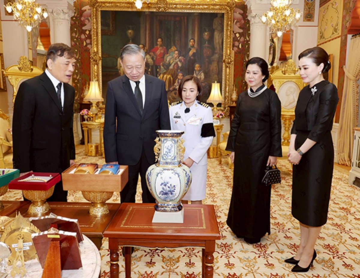 Group of five formally dressed people standing around an ornate display of gifts and a large blue-and-white porcelain vase on a wooden table in a luxurious, gold-accented room.