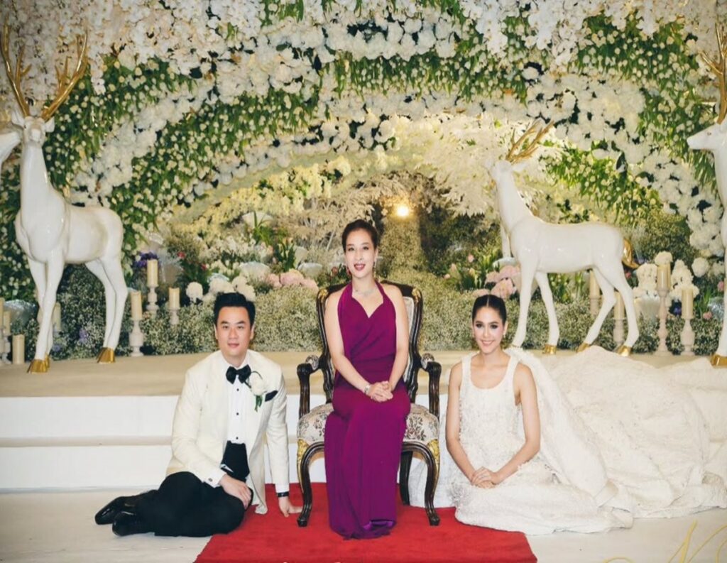 Three people pose on a red carpet in front of a white-flower wedding arch with deer statues in the background.