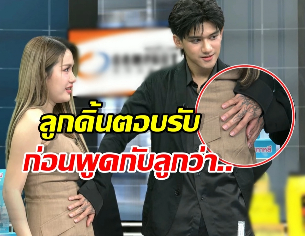 Man wearing dark clothes rests his tattooed hand on a woman's chest as they talk on a TV set; bold Thai captions overlay the image.