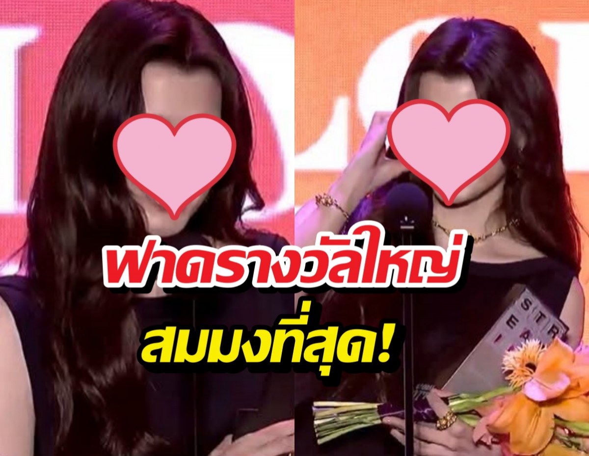 Two women on a stage at an award ceremony; pink background, pink hearts cover their faces, with bold Thai caption overlaid and a bouquet visible.