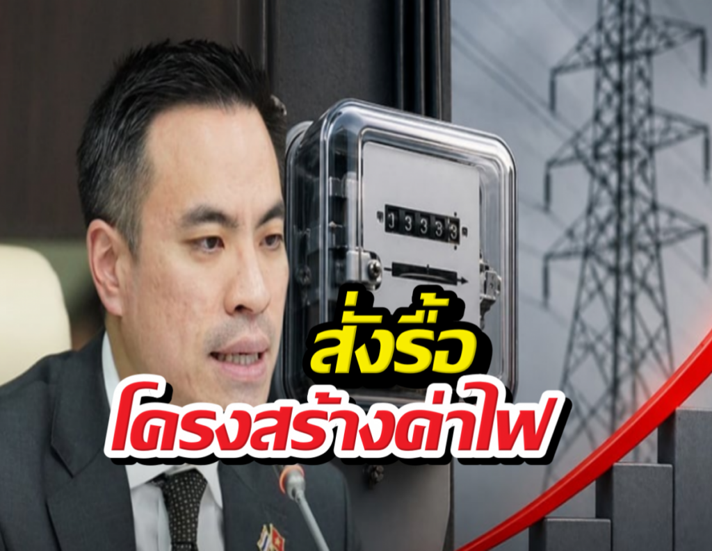 Man in a suit speaks into a microphone beside a wall-mounted safe, with power‑line towers in the background.