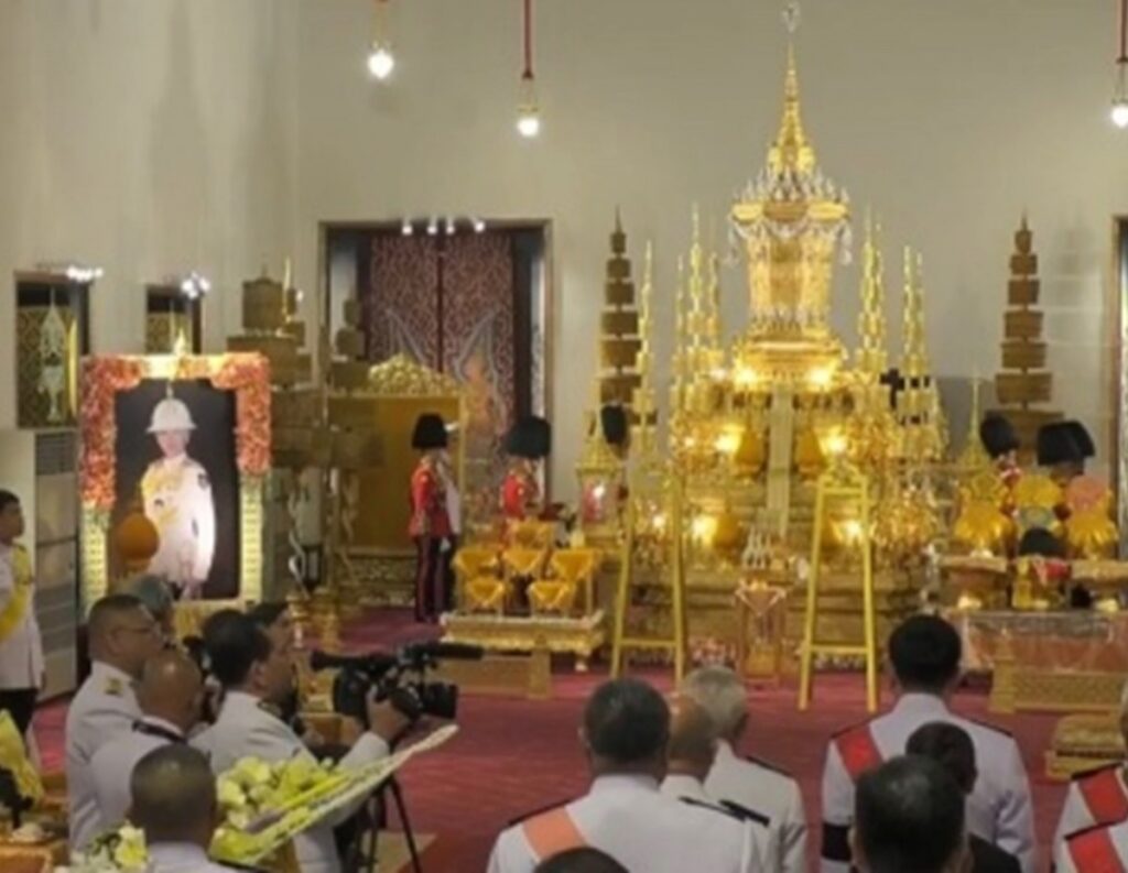 Gilded Thai ceremonial altar with tiered spires and surrounding offerings in a temple hall; attendees in white uniforms sit on a red carpet.